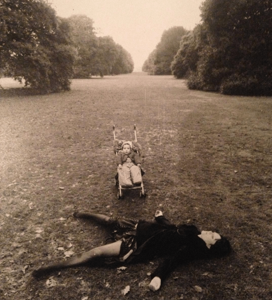 A photograph of Amy as a baby by Hag. They are in a large London park. Her mum is lying corpse like, on the grass in the foreground and Amy is in her buggy. 
